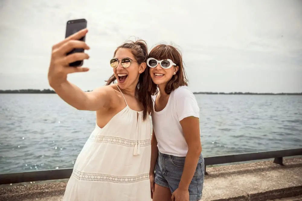 Two women smile and take a selfie beside the water with beach chairs and umbrellas reflected in their sunglasses.