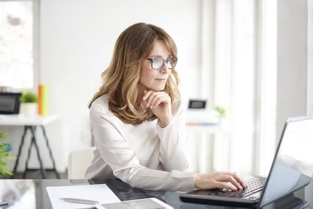 Woman with glasses wearing a white sweater looking at her computer in her home office.