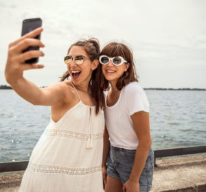 Two women smile and take a selfie beside the water with beach chairs and umbrellas reflected in their sunglasses.