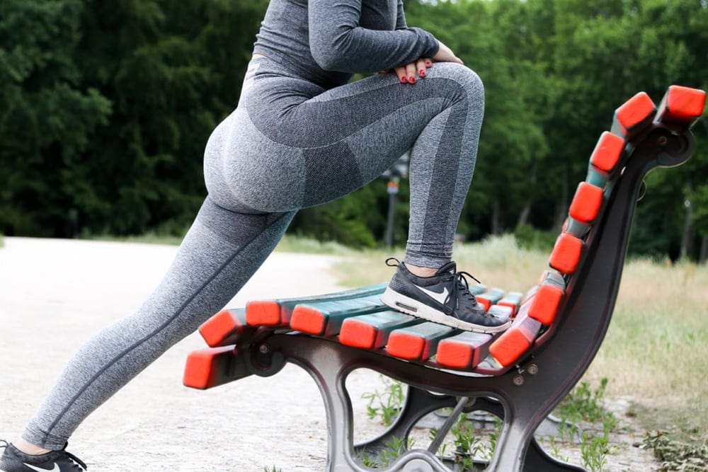 Woman in running tights doing a one legged stretch using a park bench.